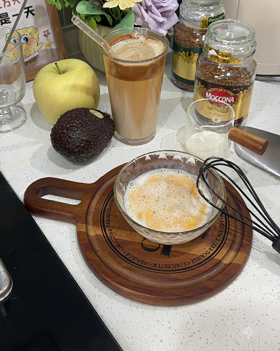 A glass bowl filled with a beaten frothy mixture of egg and milk next to a black whisk.