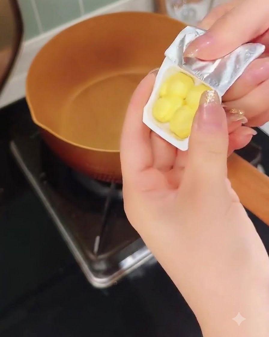 A hand peeling a small plastic container of yellow butter pieces above a warm copper pan.