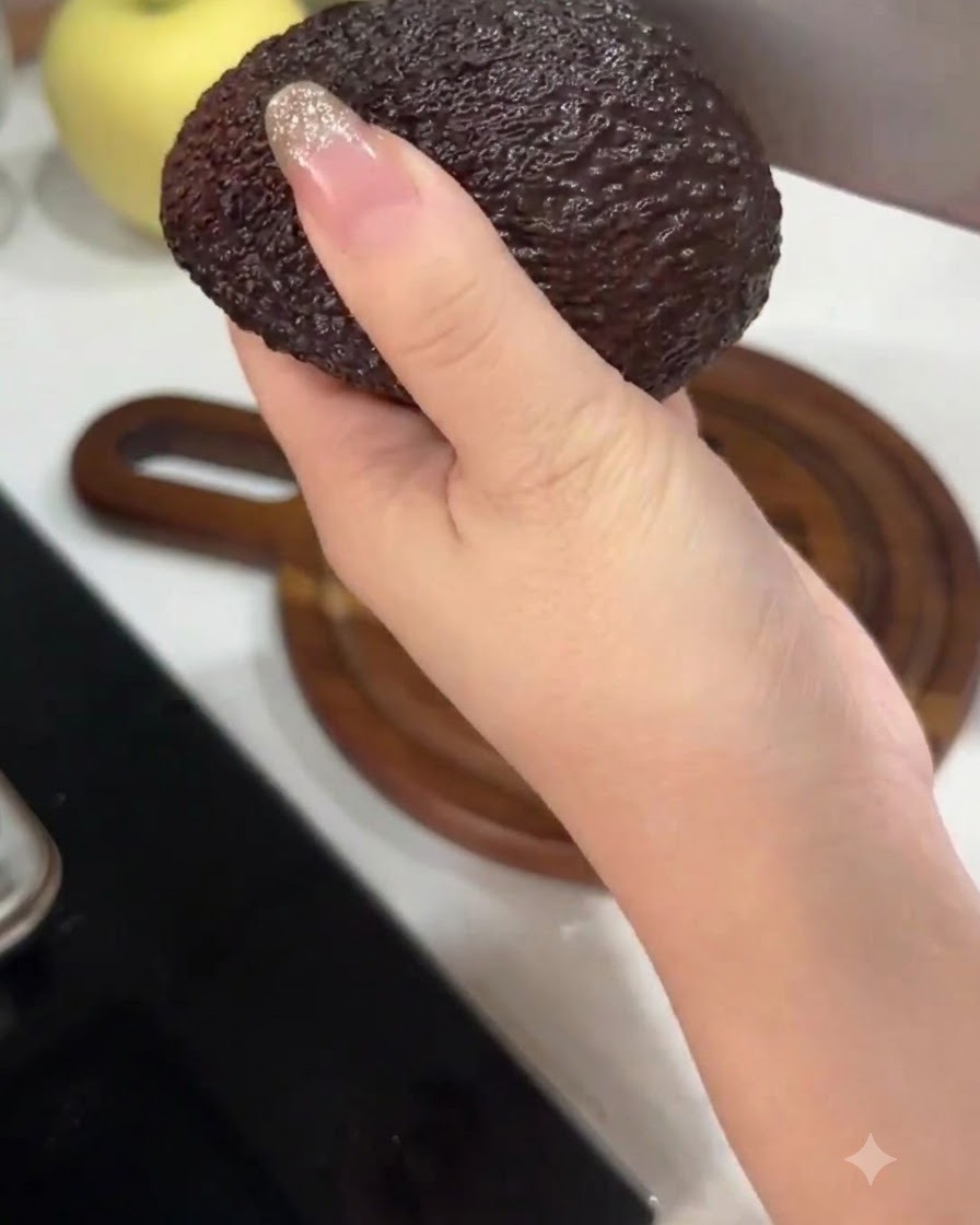 A hand holding an avocado while cutting it in half with a large kitchen knife.