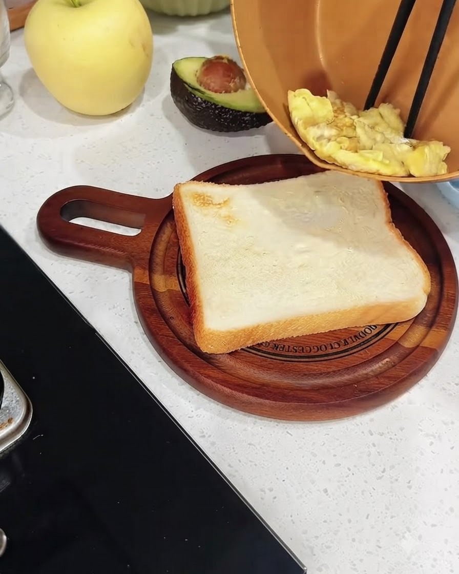 Cooked scrambled eggs being transferred from a copper pan onto a slice of toasted white bread on a wooden board.