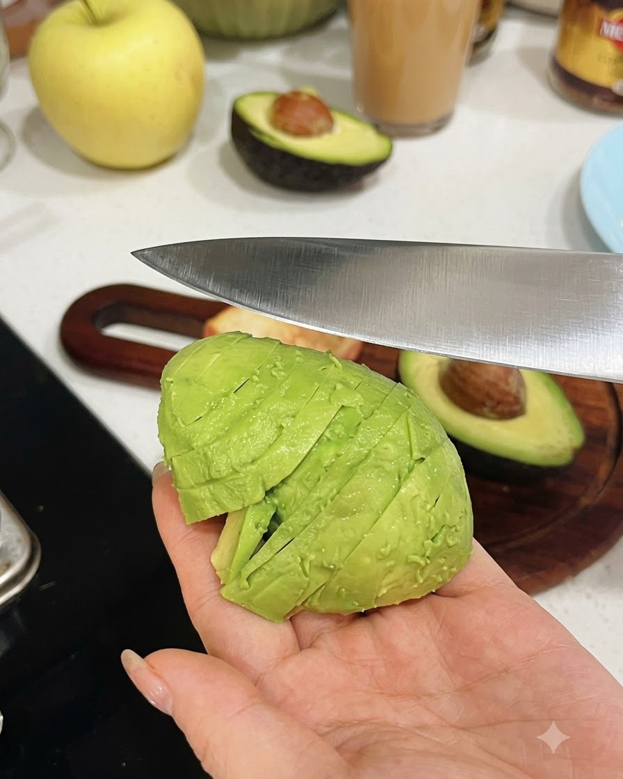 A hand holding a peeled avocado half while a kitchen knife slices it into uniform pieces.