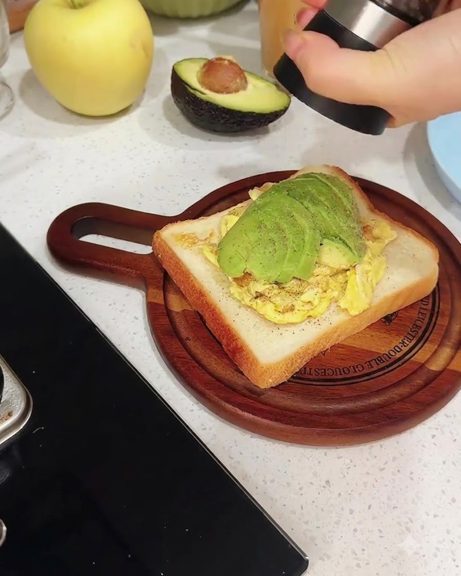 A pepper mill grinding fresh black pepper directly over a slice of toast layered with scrambled eggs and sliced avocado.