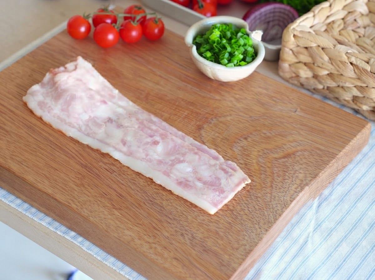A single raw bacon strip laid out on a wooden cutting board ready to be sliced.