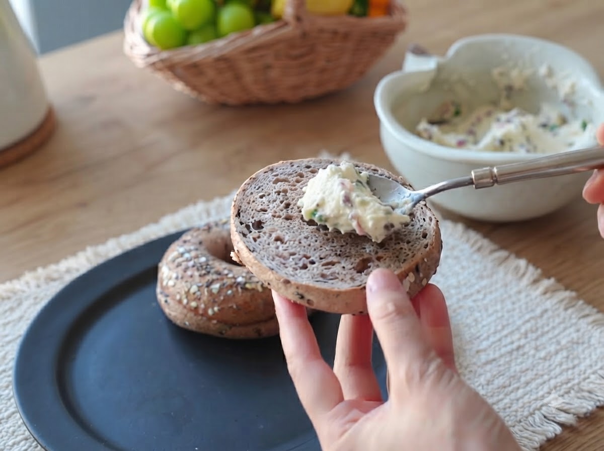 A spoonful of creamy bacon and scallion spread being smoothed onto the surface of a bagel half.