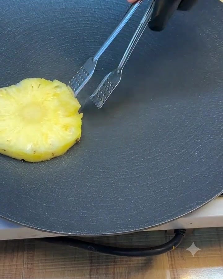 A single pineapple ring being turned on a dark griddle using metal tongs.