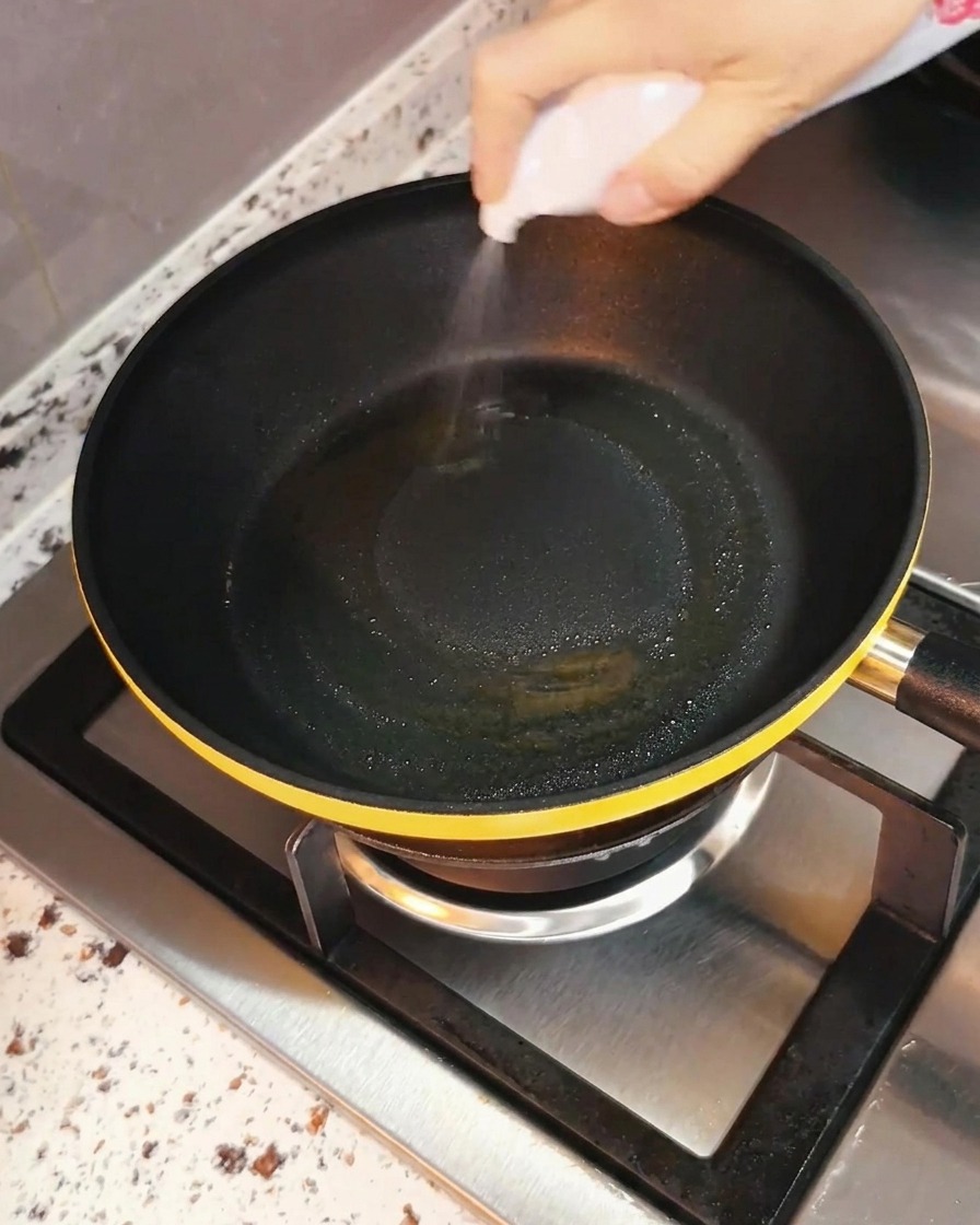 A hand spraying cooking oil evenly into a black frying pan set over a stove burner.