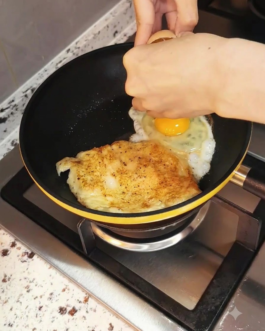 An egg being cracked into the frying pan beside the cooking chicken breast.