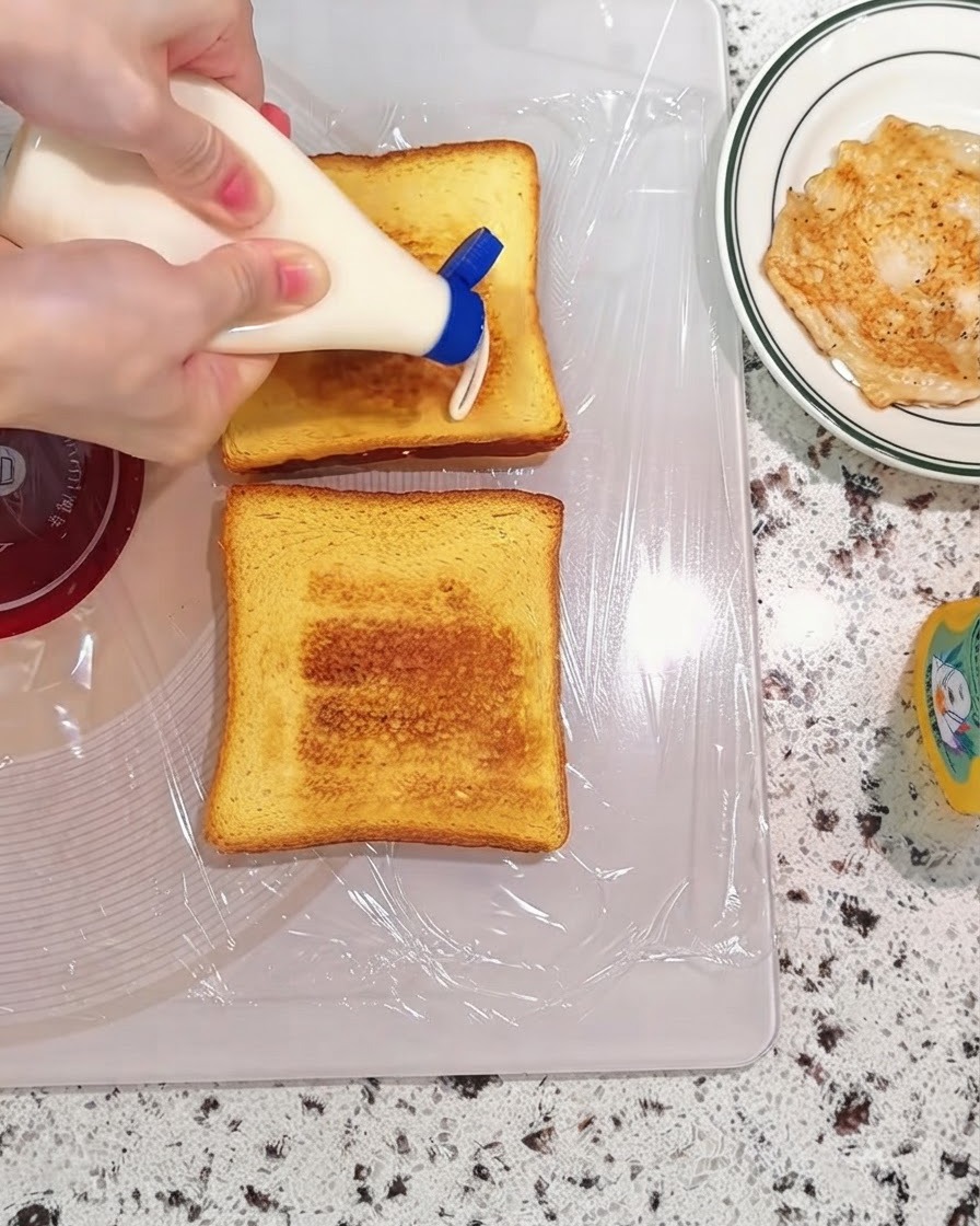 Mayonnaise being squeezed from a bottle onto toasted bread slices on a cutting board.