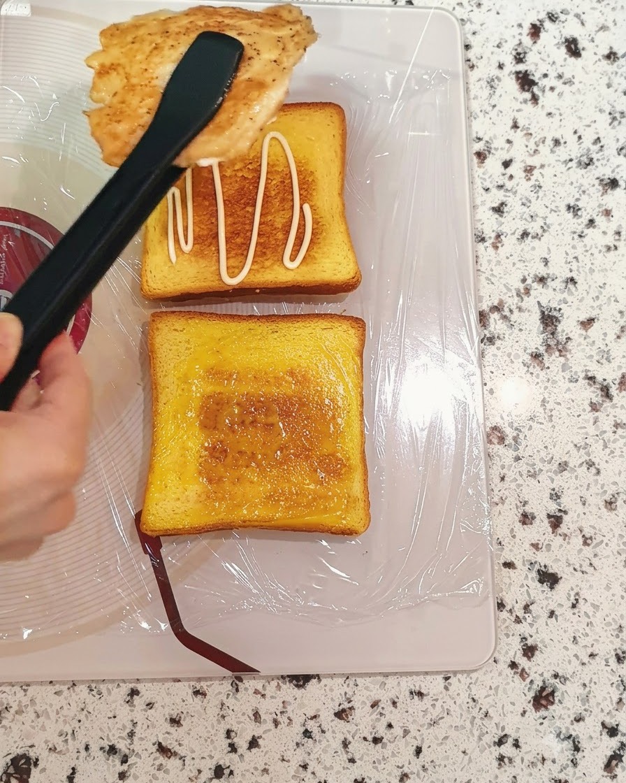 Cooked chicken breast being transferred with tongs onto a slice of sauce-spread toast.