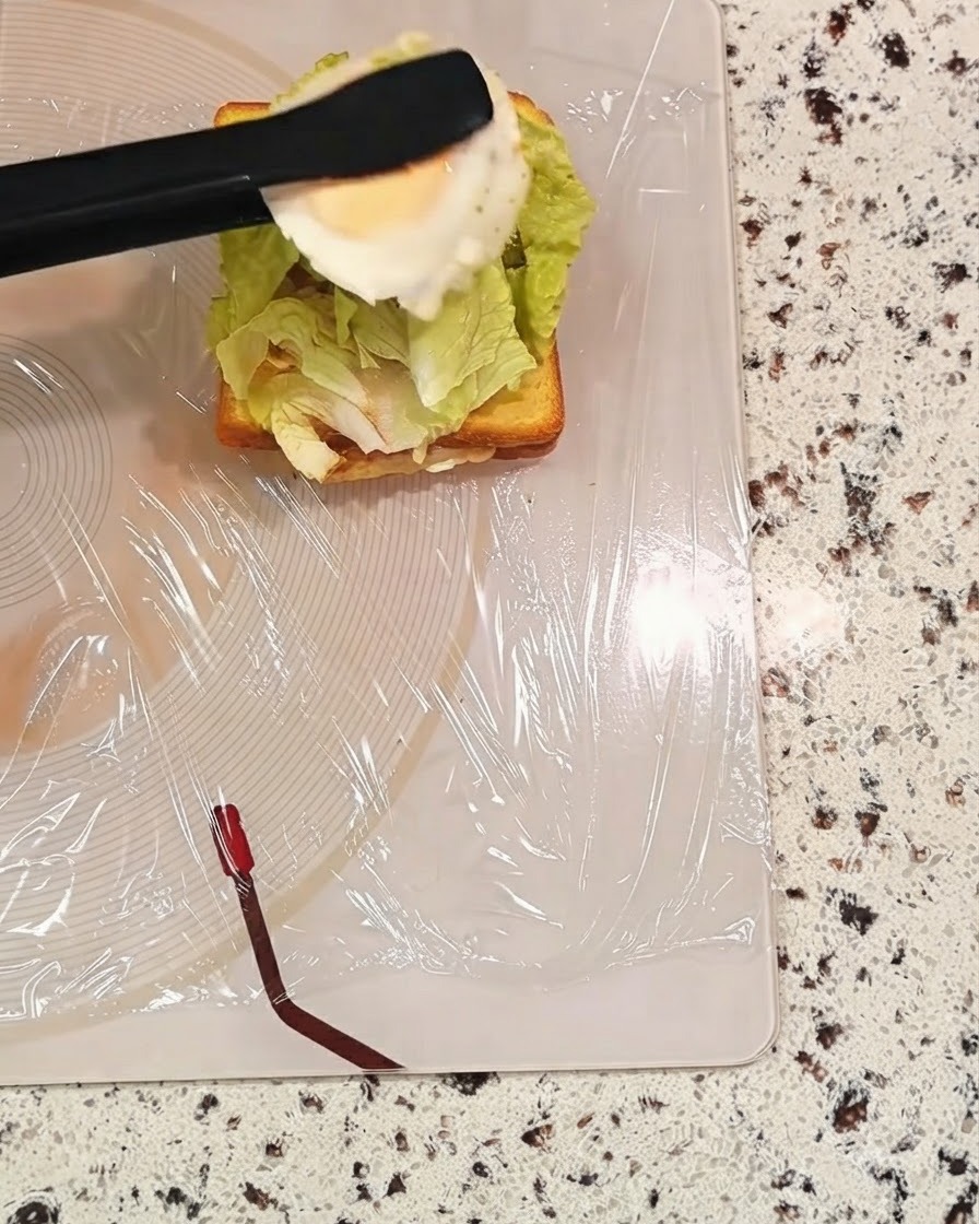 A pair of black tongs holding a cooked egg over a sandwich layer with lettuce.