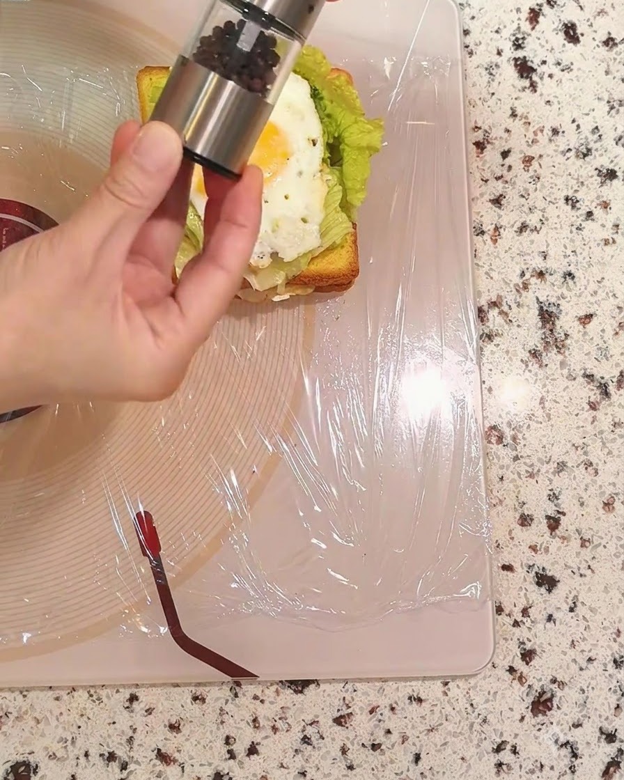 A hand holding a pepper grinder and sprinkling black pepper over the fried egg.