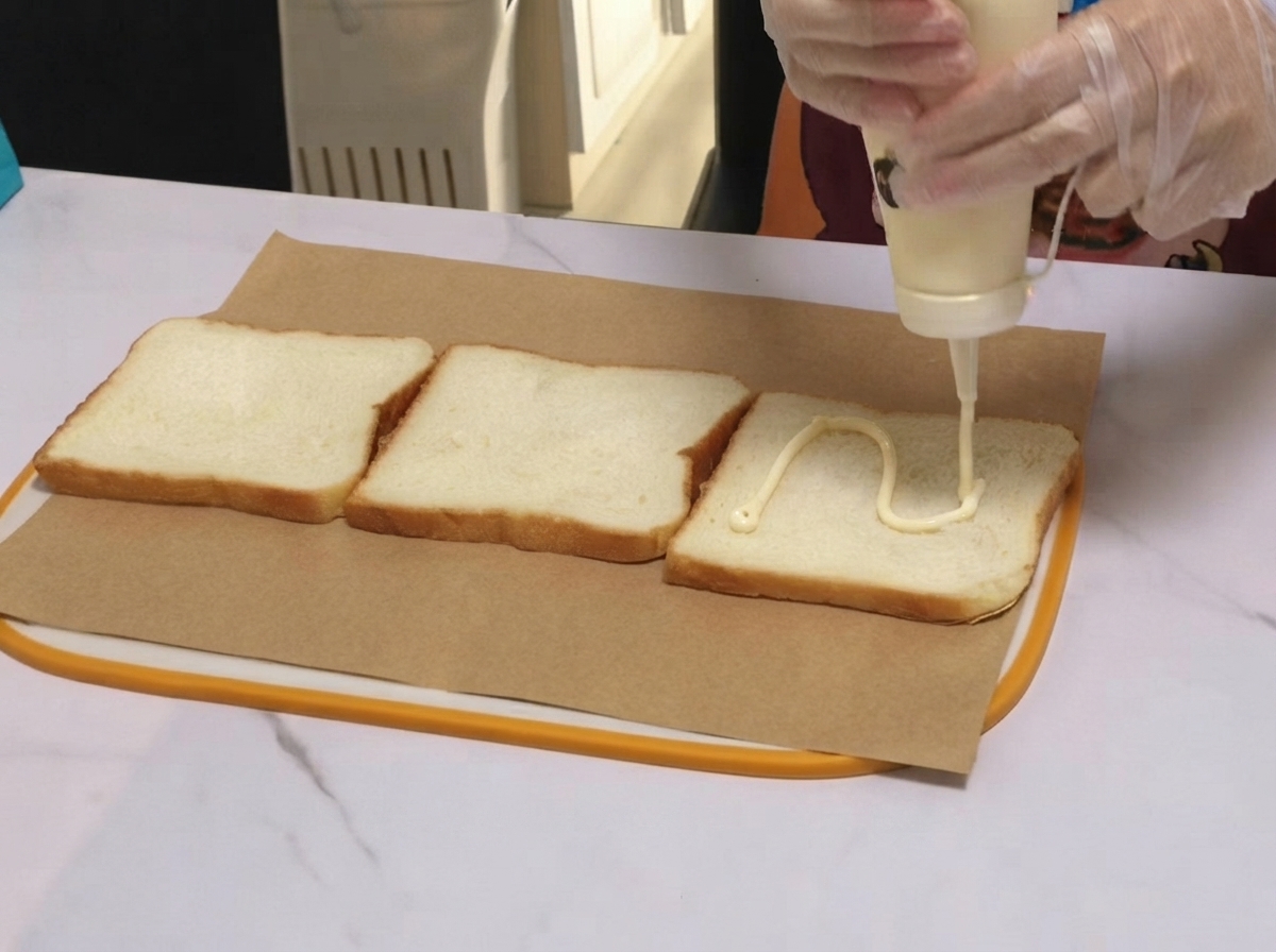 White dressing being piped in a wavy line across a piece of sandwich bread on parchment paper.