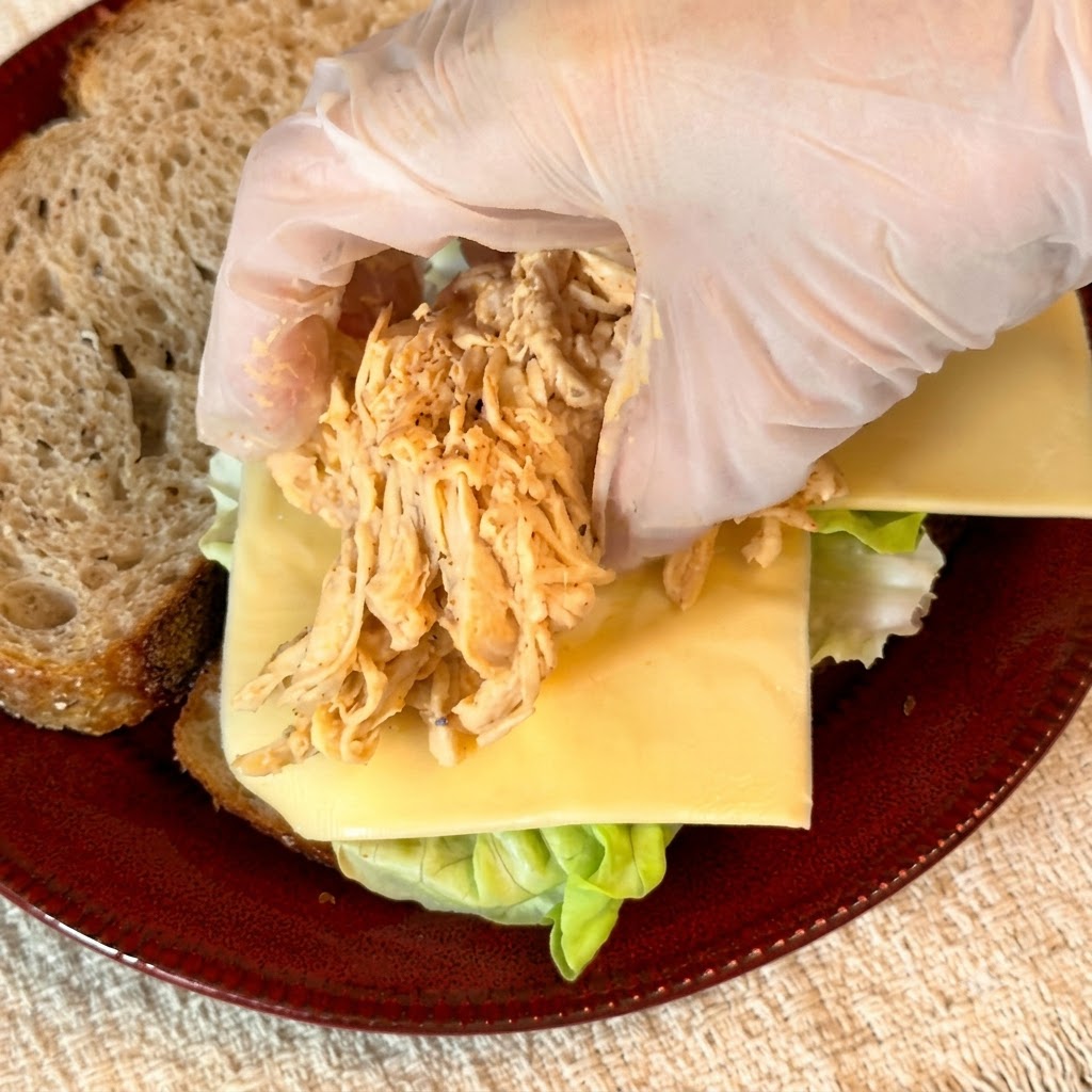 Gloves placing a mound of seasoned shredded chicken onto the cheese layer of the sandwich.