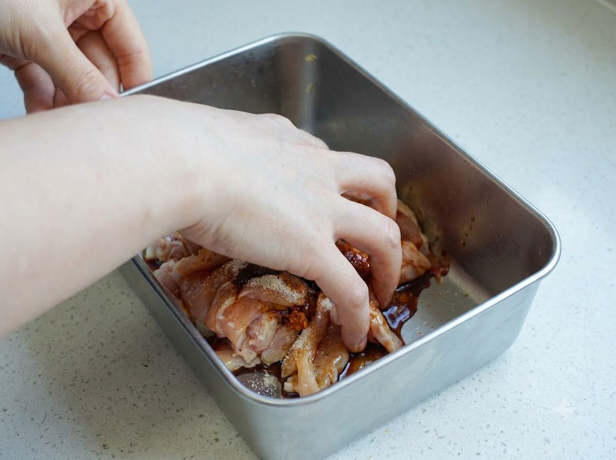 A hand mixing raw seasoned chicken slices inside a metal container.
