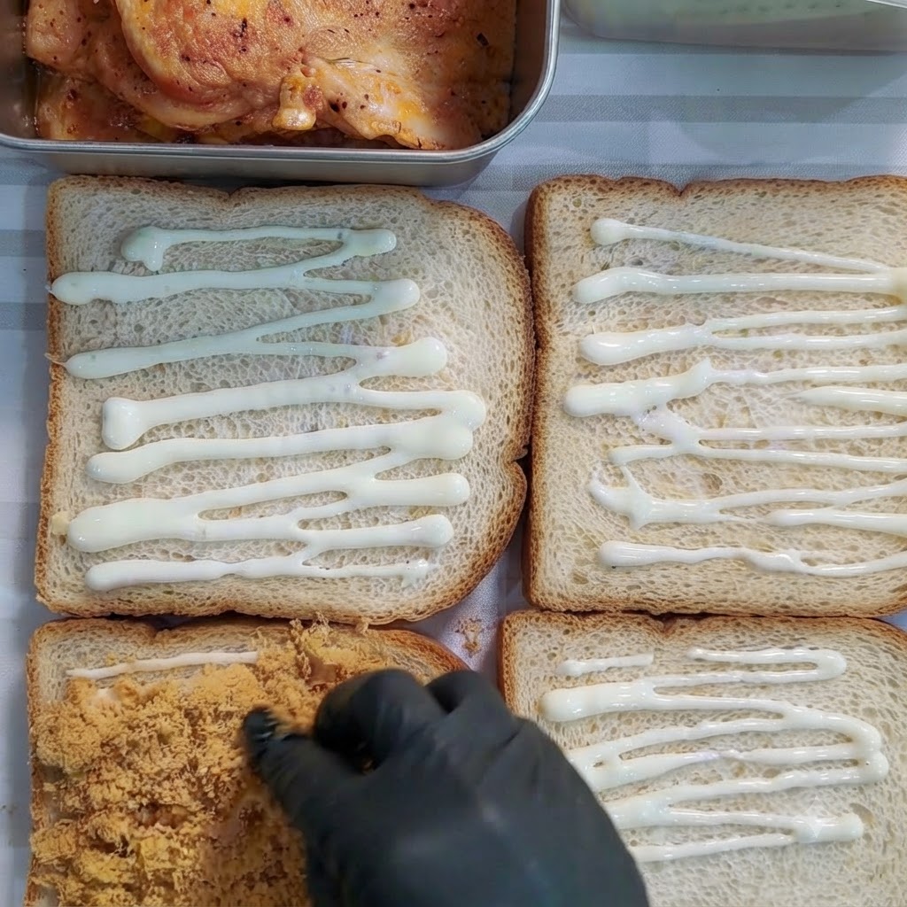 Gloved hand spreading golden-brown meat floss onto a slice of bread covered with dressing.