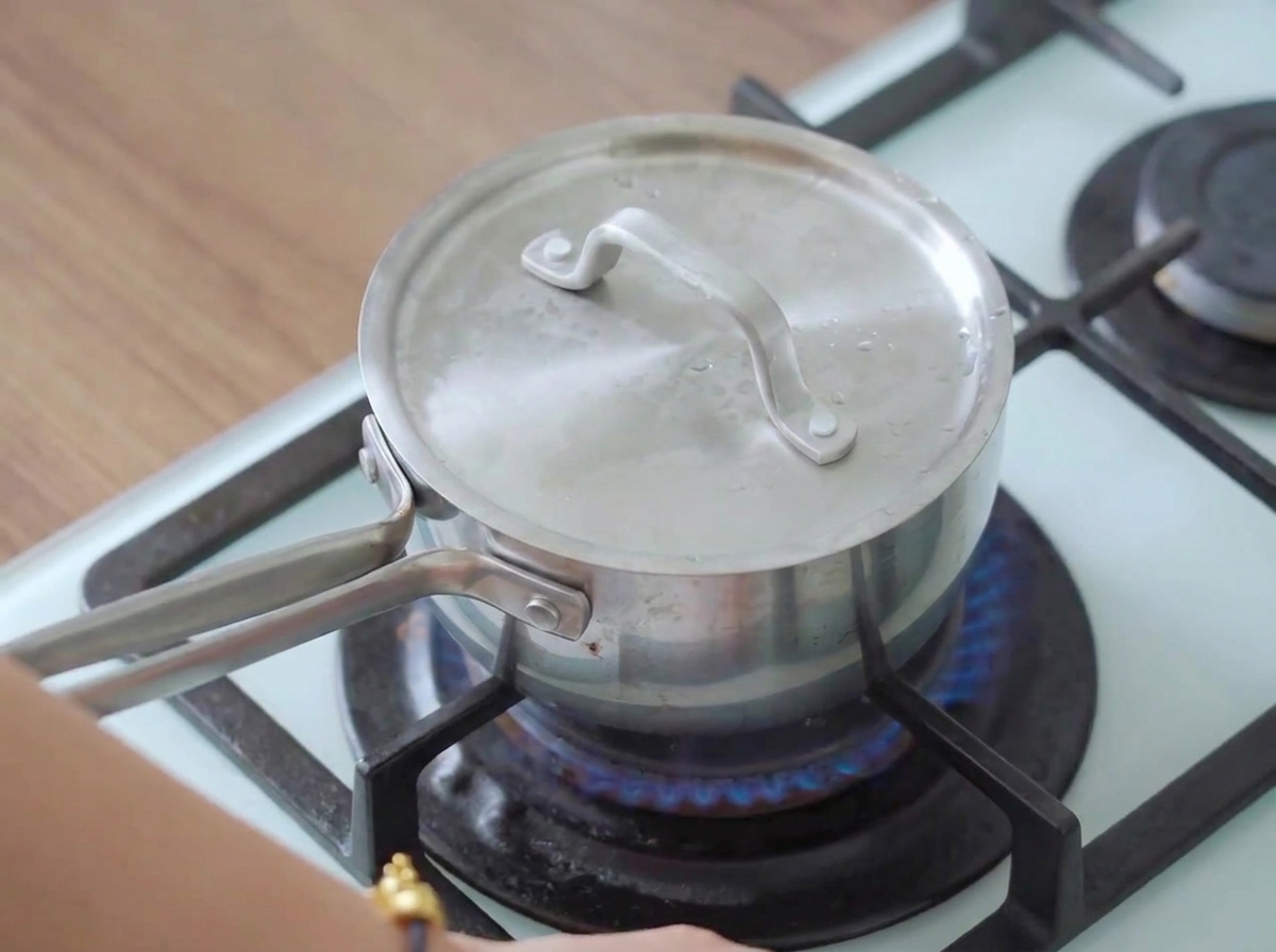 A covered pot on a lit stovetop with steam escaping, cooking the chicken breast.