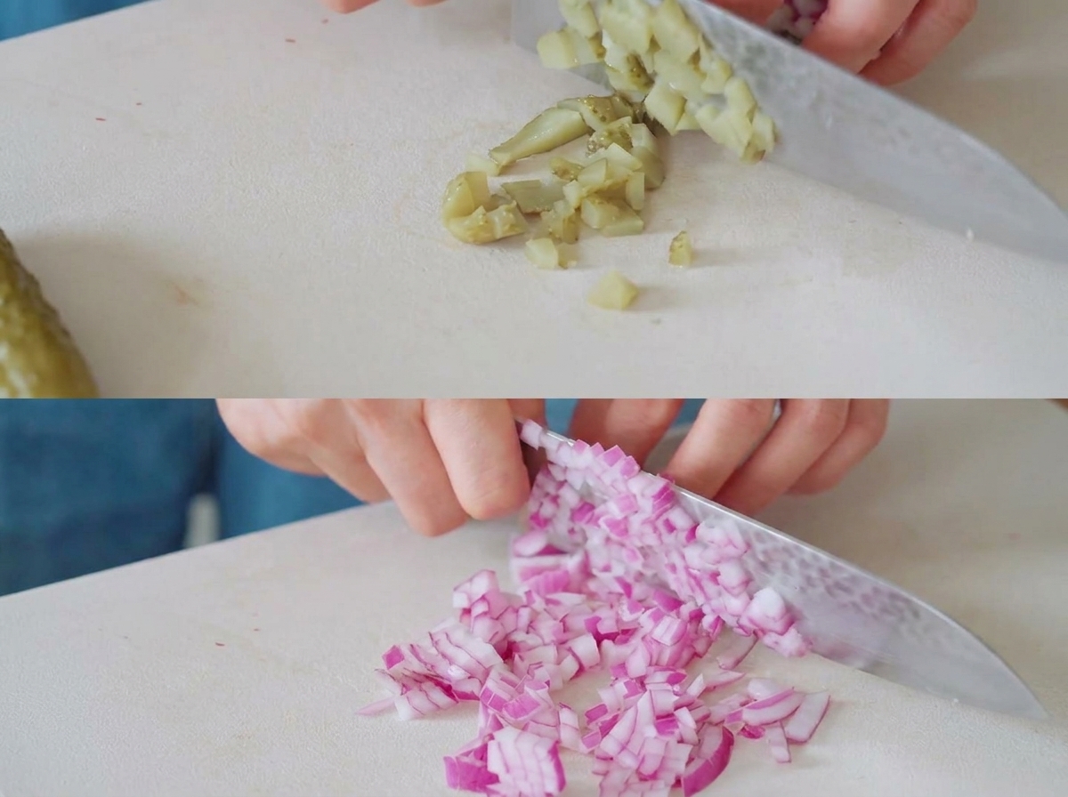 Pickles and red onions being chopped into small pieces on a white cutting board with a chefs knife.
