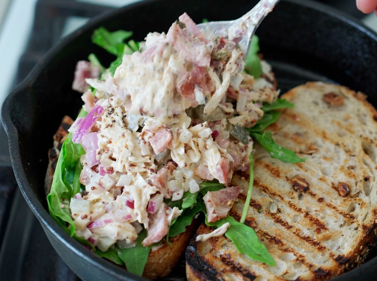 A spoonful of shredded chicken salad being placed onto fresh green arugula on a sourdough sandwich slice.