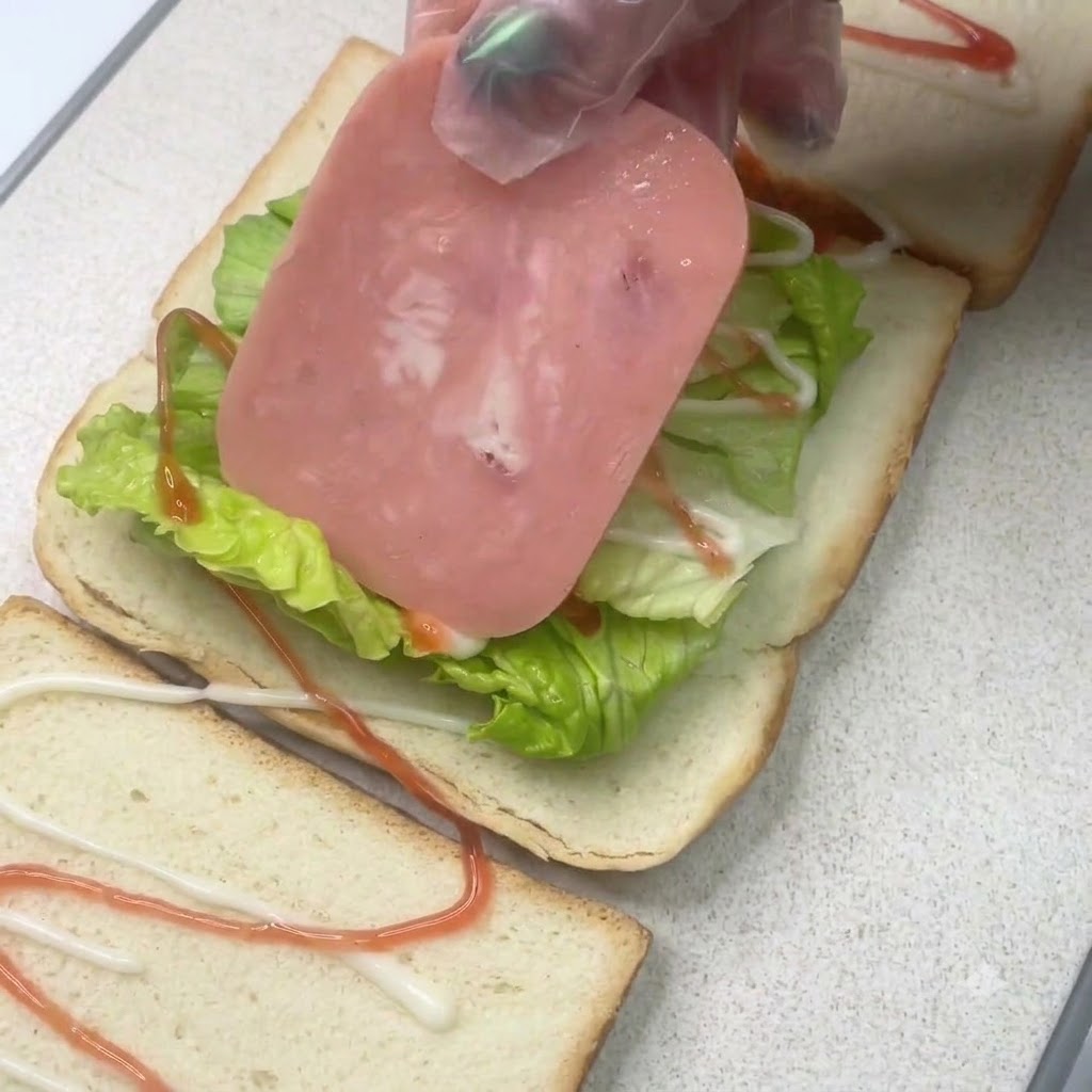 A slice of ham being placed on top of a lettuce layer on a piece of sandwich bread with dressings.