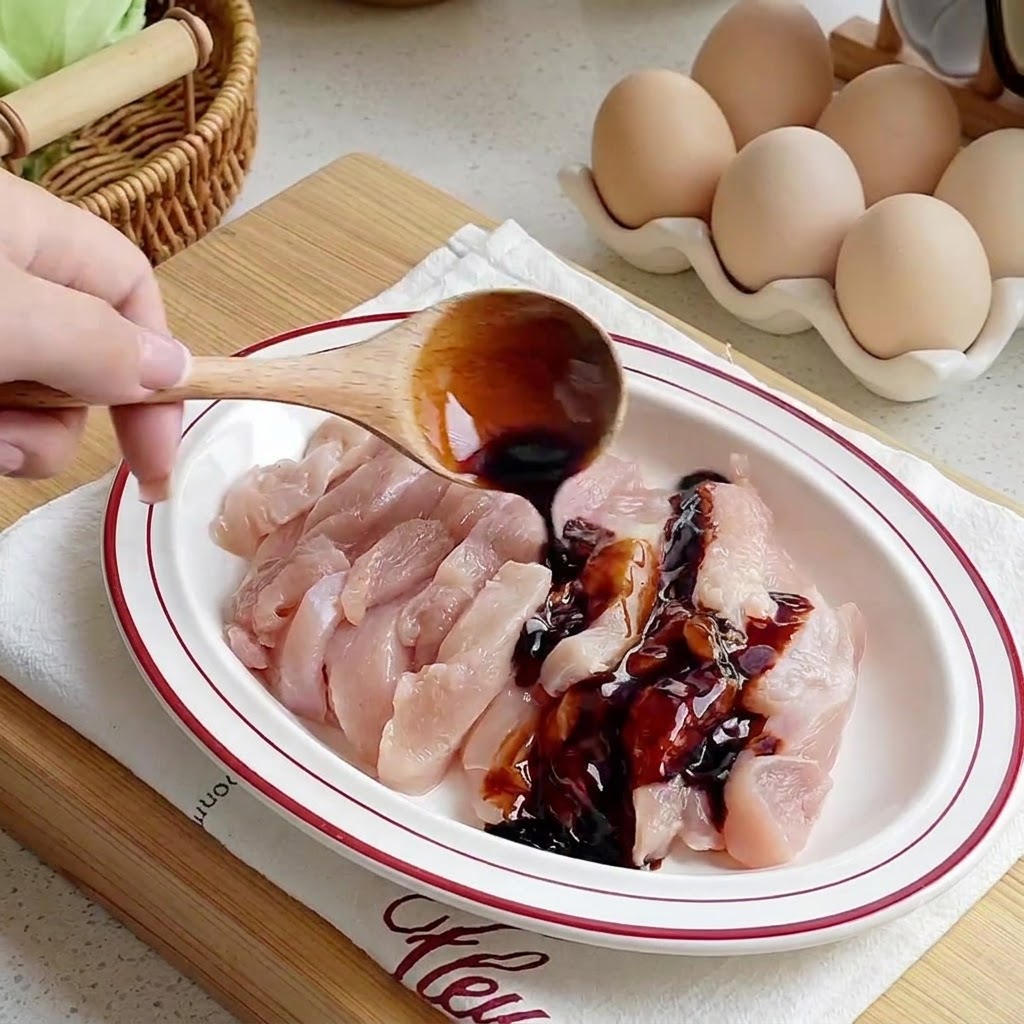 Sauces being poured from a wooden spoon onto raw chicken strips arranged in a white plate.