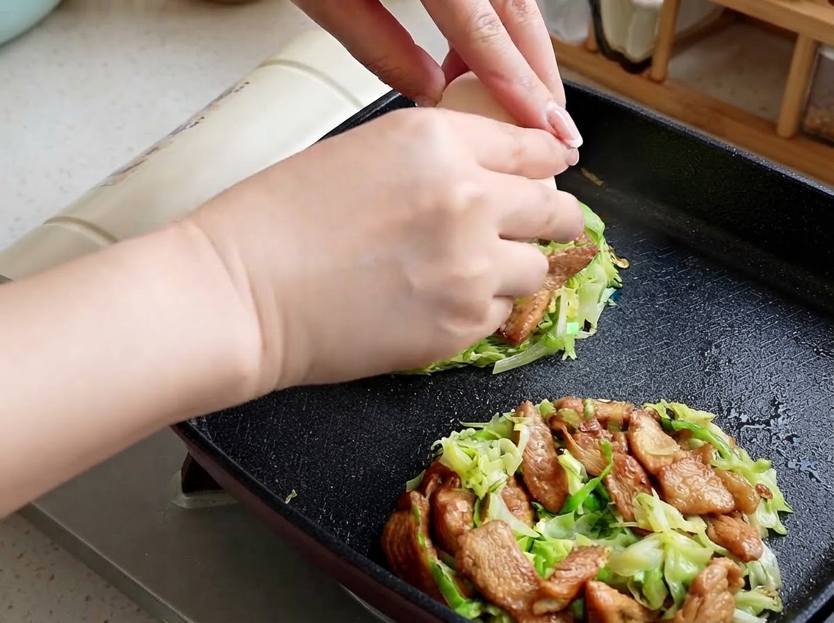 Hands cracking a raw egg onto the center of a cabbage and chicken portion in a pan.