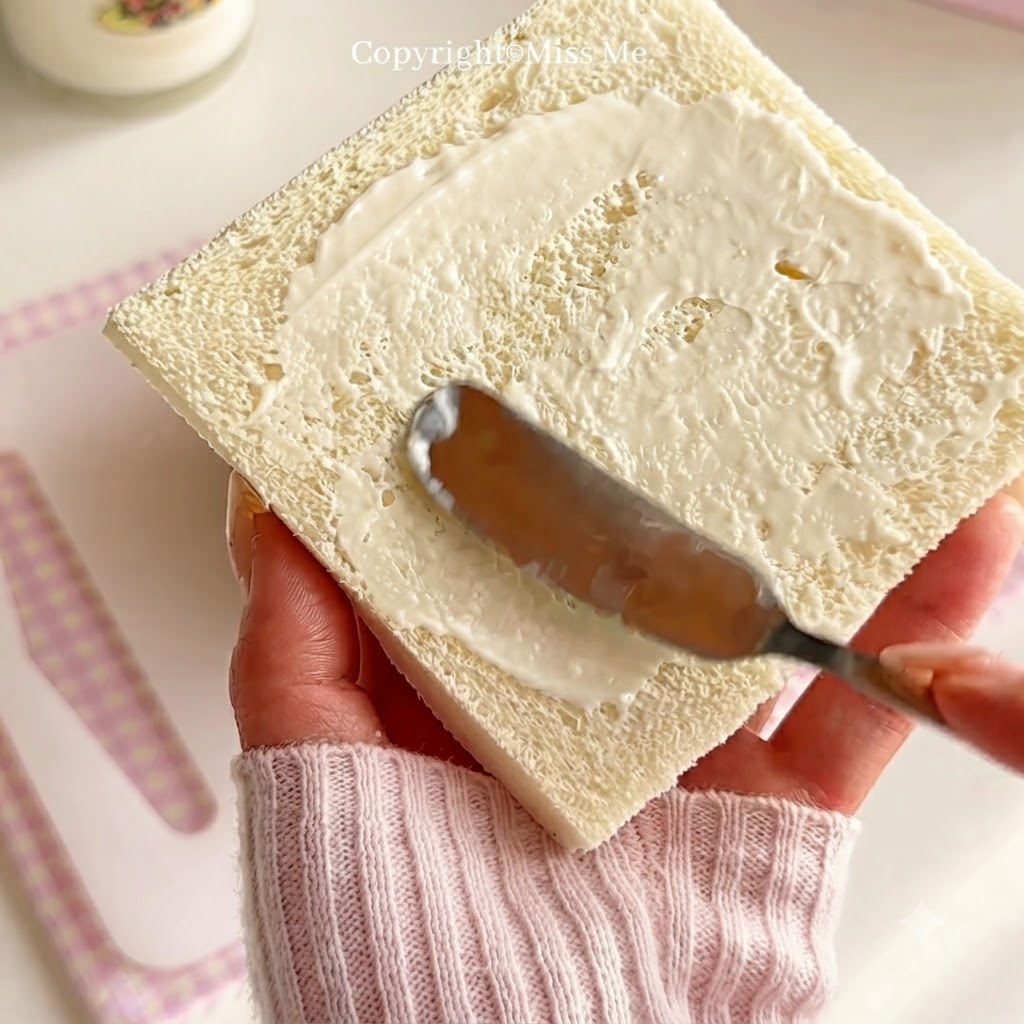 A hand holding a knife to spread white salad dressing evenly across a square slice of white bread.