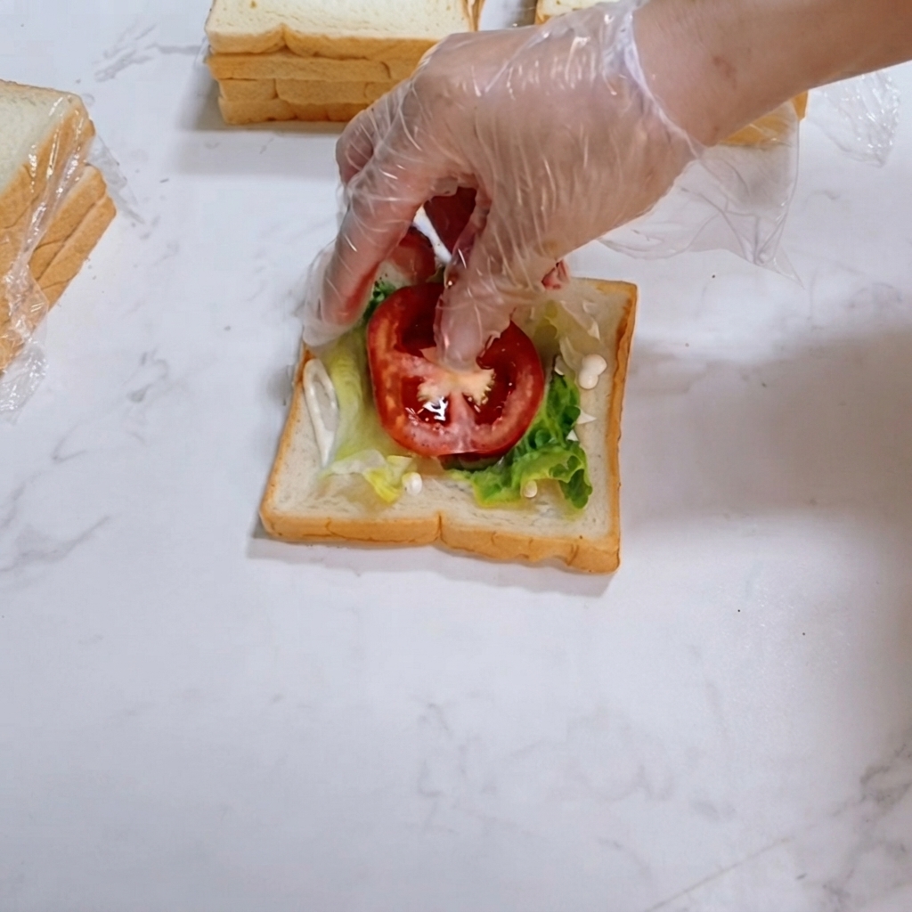 Fresh red tomato slices being carefully laid on top of green lettuce on the bread slice.