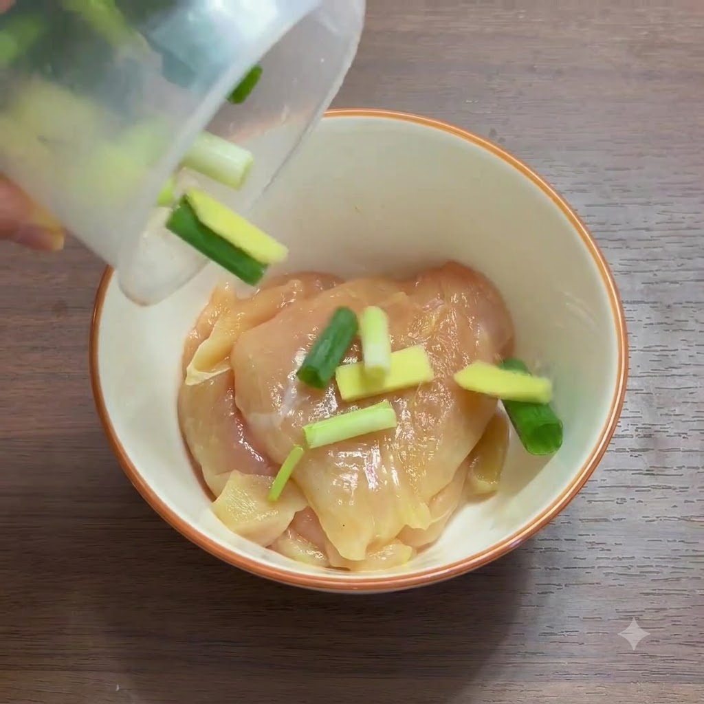 A close-up of sliced chicken breast in a bowl being mixed with scallions and ginger slices.