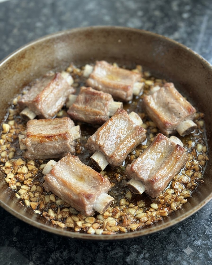 Blanch pork ribs browning in a pan surrounded by fried garlic and oil.