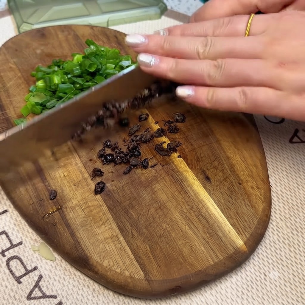 Fermented black beans and chopped green onions being minced on a wooden cutting board with a knife.