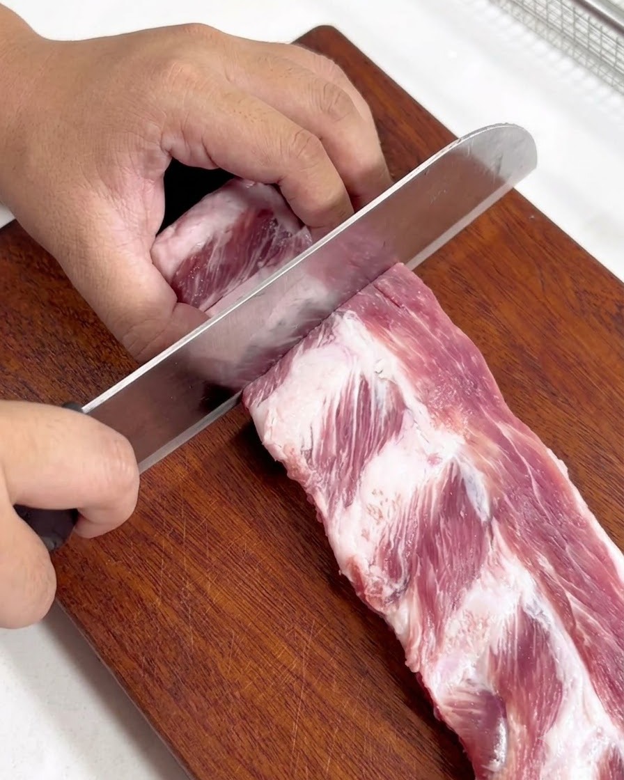 A rack of raw pork ribs being cut into smaller individual pieces on a wooden cutting board.