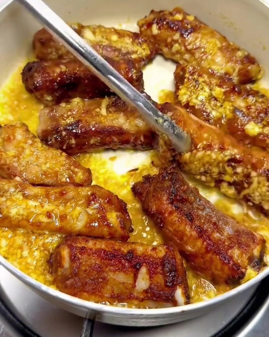 Pork ribs being turned with metal tongs inside a pan, coated with garlic butter sauce.