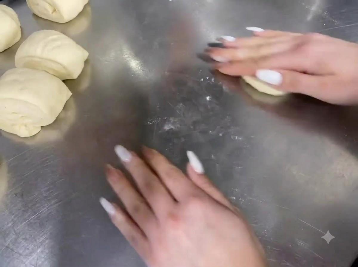 Hands rolling portions of dough on a stainless steel countertop.