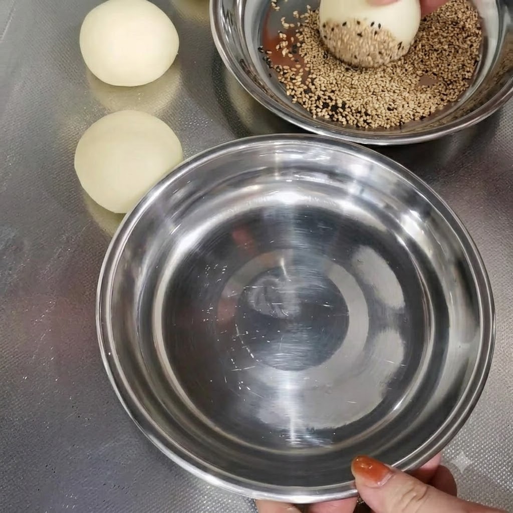 A dough ball being coated with sesame seeds in a metal bowl next to other prepared dough balls.