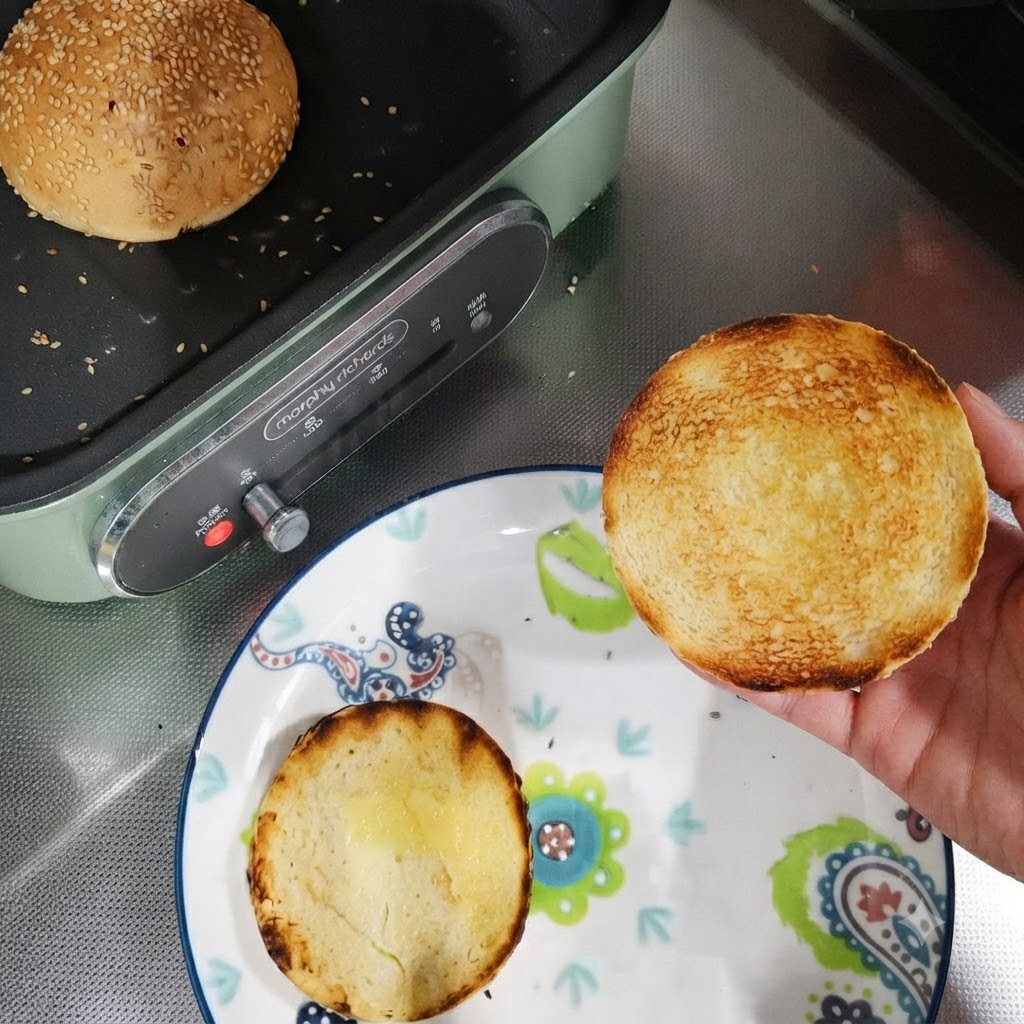 Toasted burger bun halves on a plate next to a pan with a whole bun being toasted.