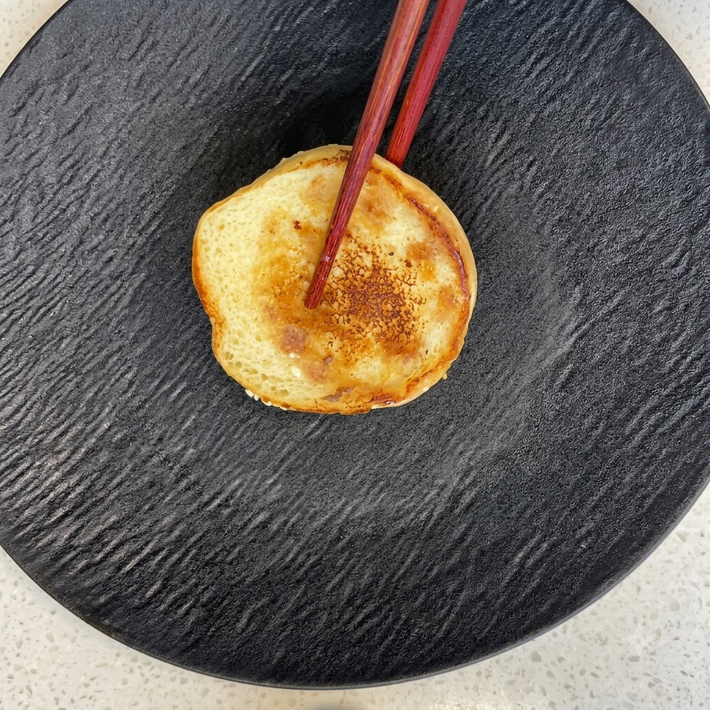 A toasted burger bun half resting on a black textured plate with chopsticks.
