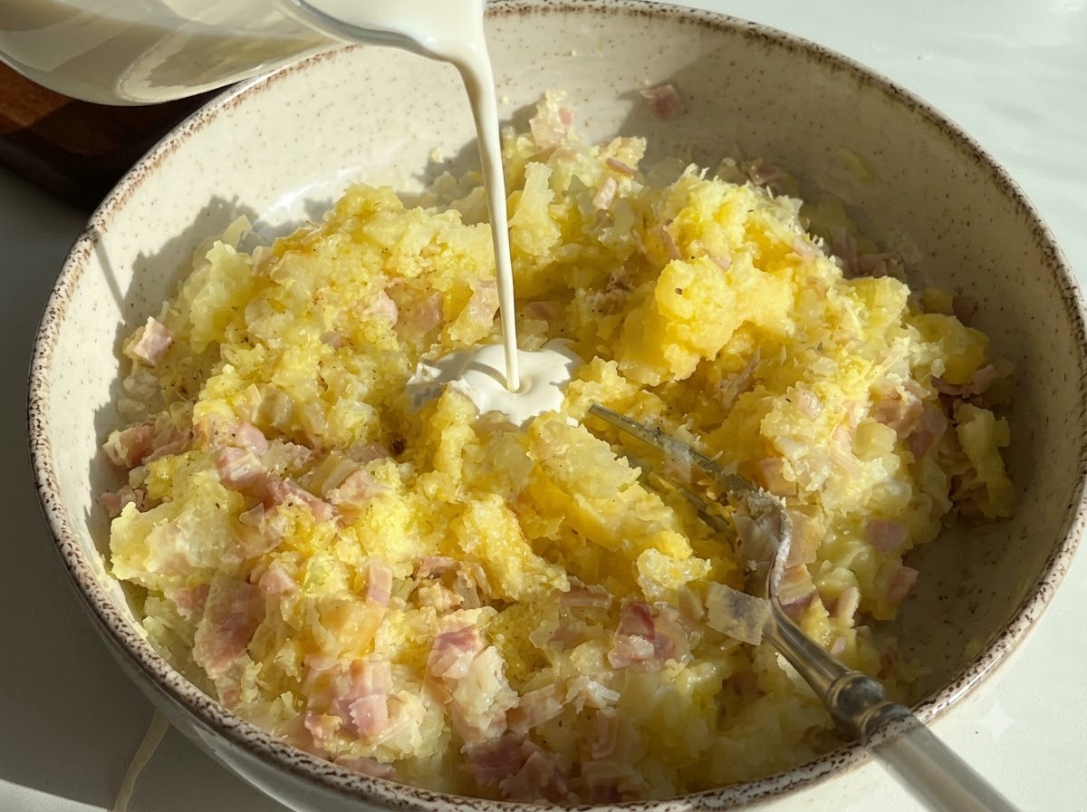 Heavy cream being poured into a bowl containing mashed potatoes and diced ham.