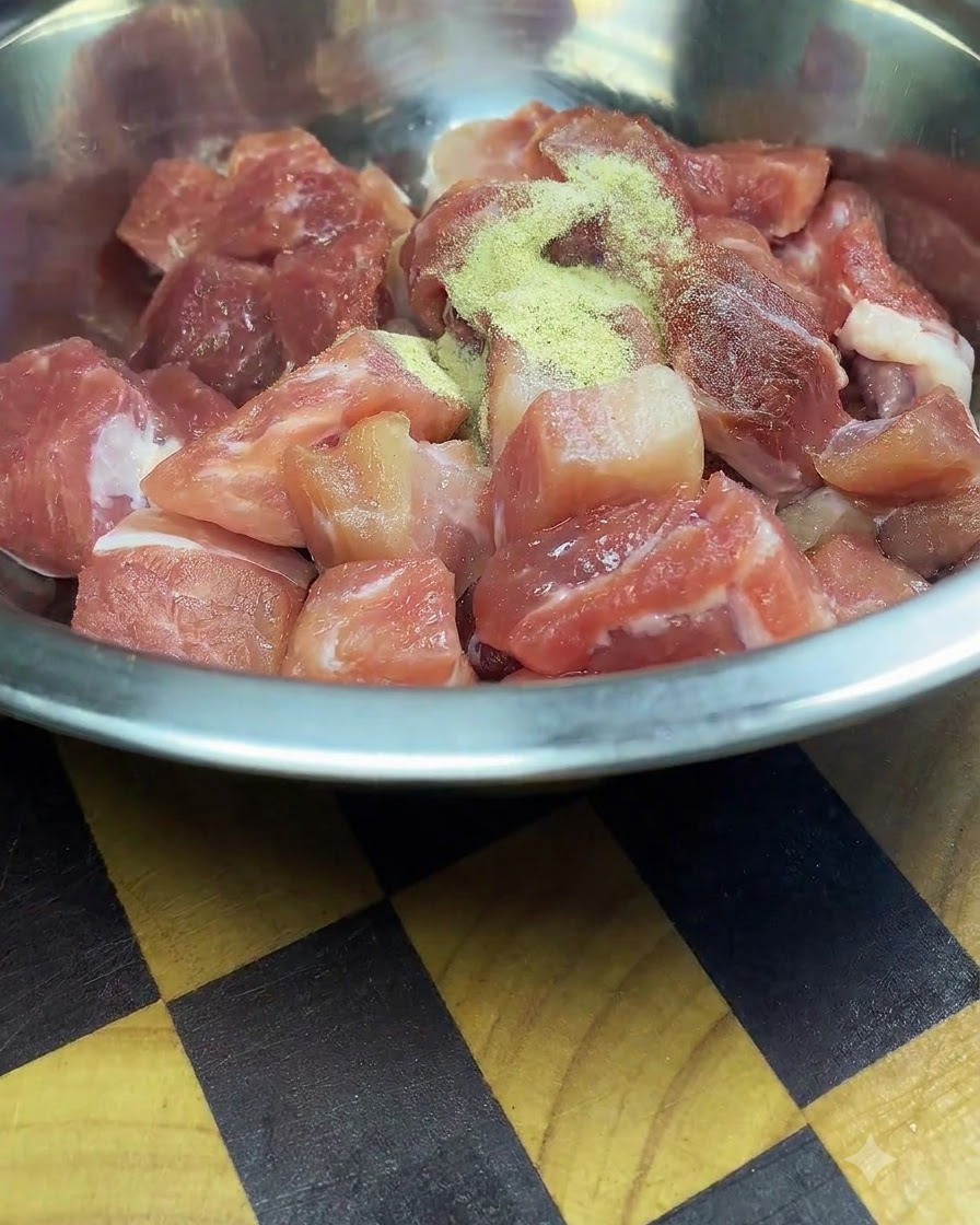 Freshly cut raw pork cubes inside a stainless steel mixing bowl sprinkled with seasonings and cornstarch.