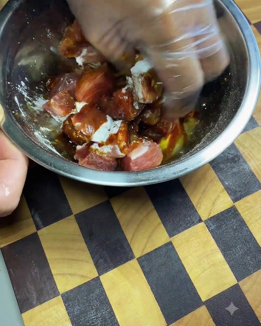 A gloved hand kneading and massaging dark soy sauce and spices into pork cubes inside a metal bowl.