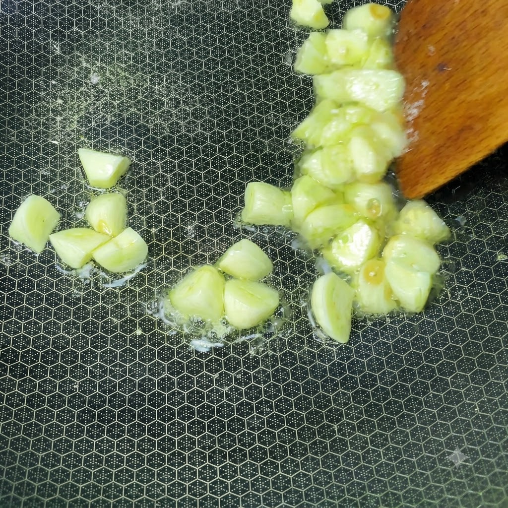 Freshly chopped garlic cloves sizzling in hot oil inside a wok being stirred with a wooden spatula.