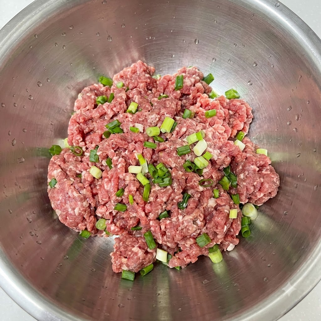 Chopped green onions freshly placed on top of ground meat in a stainless steel bowl.