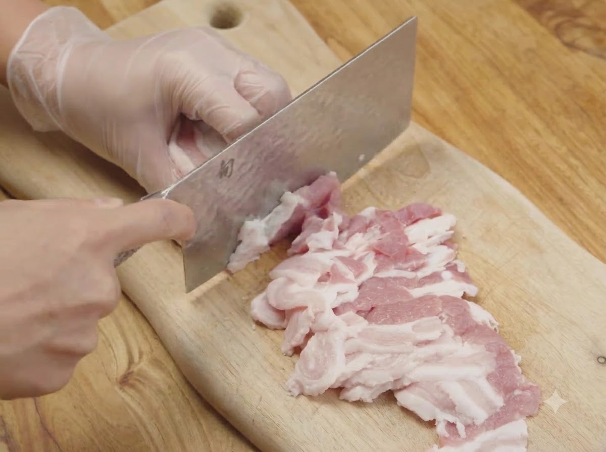 Raw pork belly being sliced into thin strips on a wooden cutting board using a kitchen cleaver.