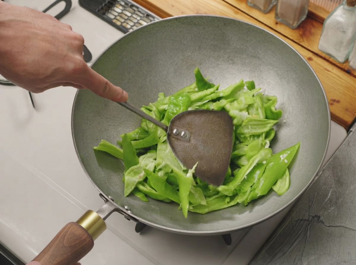 Cut green peppers being tossed with a metal spatula in a hot, dry wok.