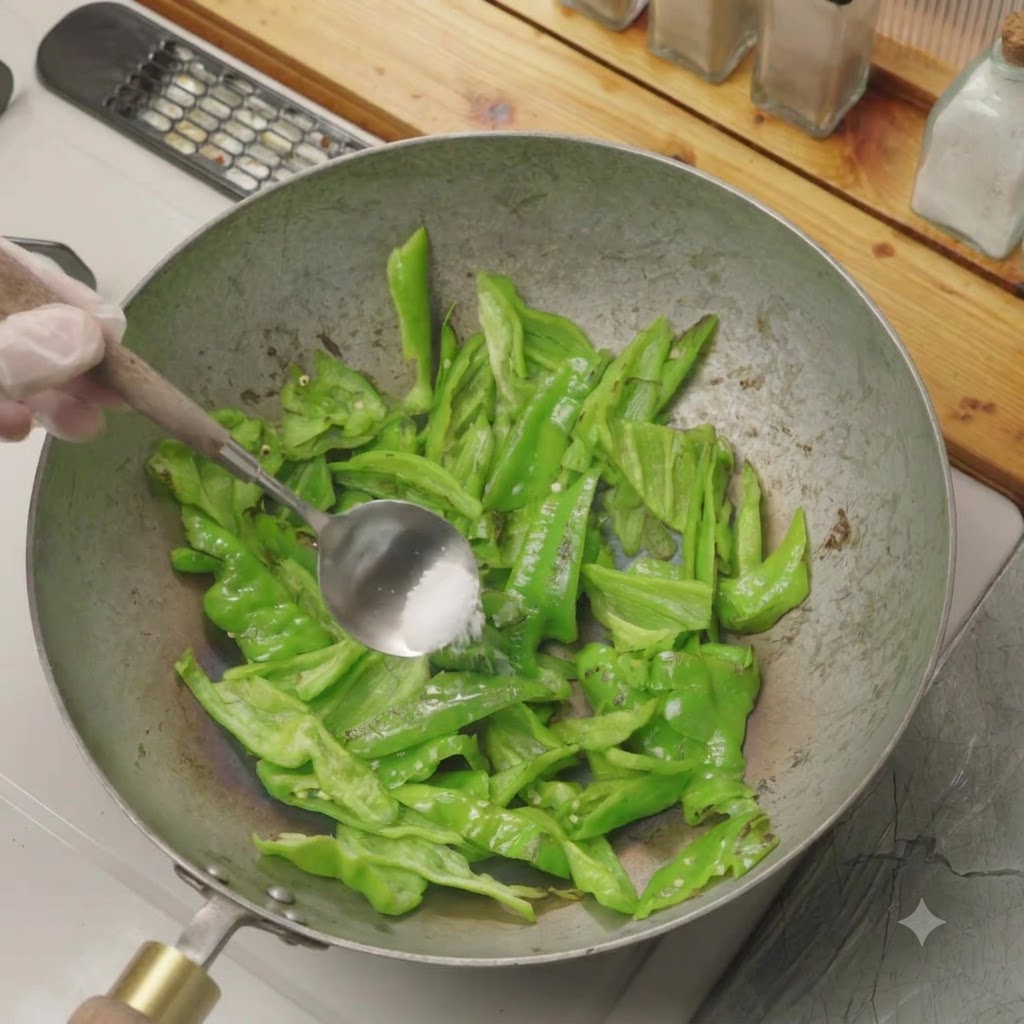 A spoonful of salt being added to sliced green peppers dry-frying in a wok.