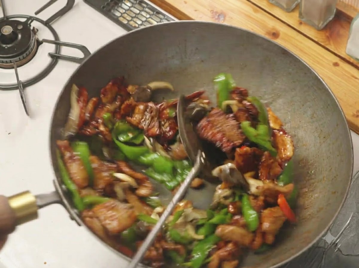 A spatula mixing green peppers, mushrooms, and pork belly together in a hot wok.