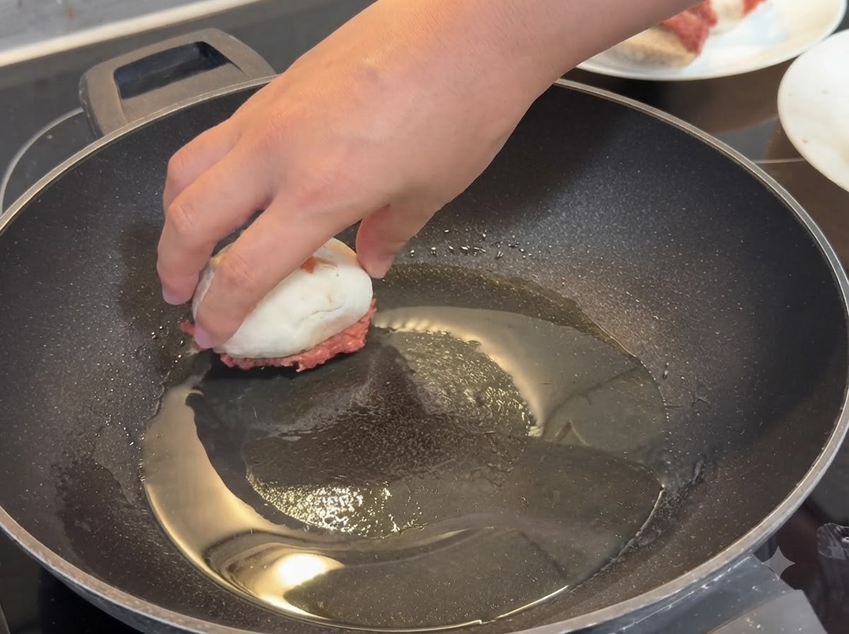 A hand placing a stuffed mushroom meat-side down into a hot frying pan coated with oil.