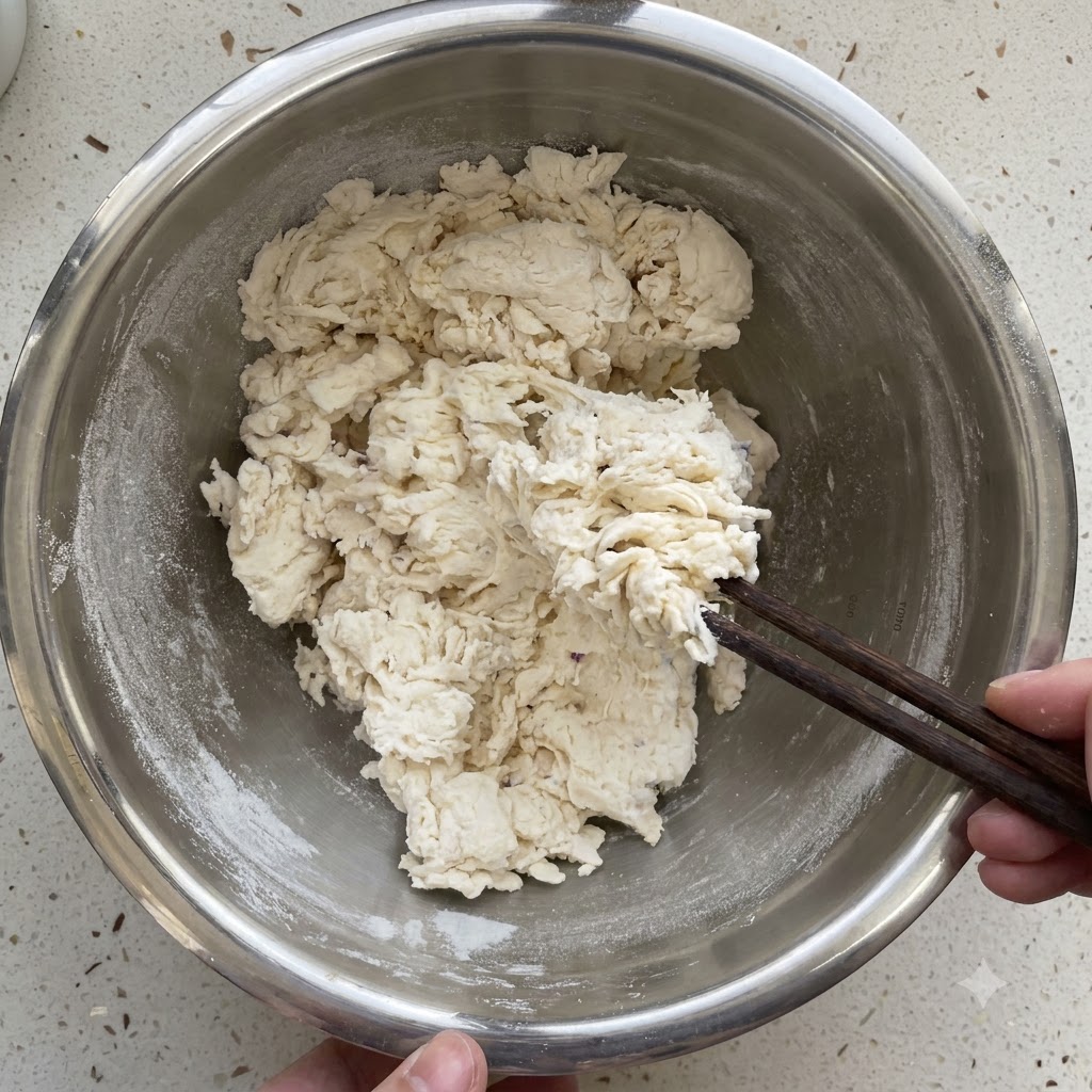 Shaggy dough being mixed and kneaded with chopsticks inside a metal bowl.
