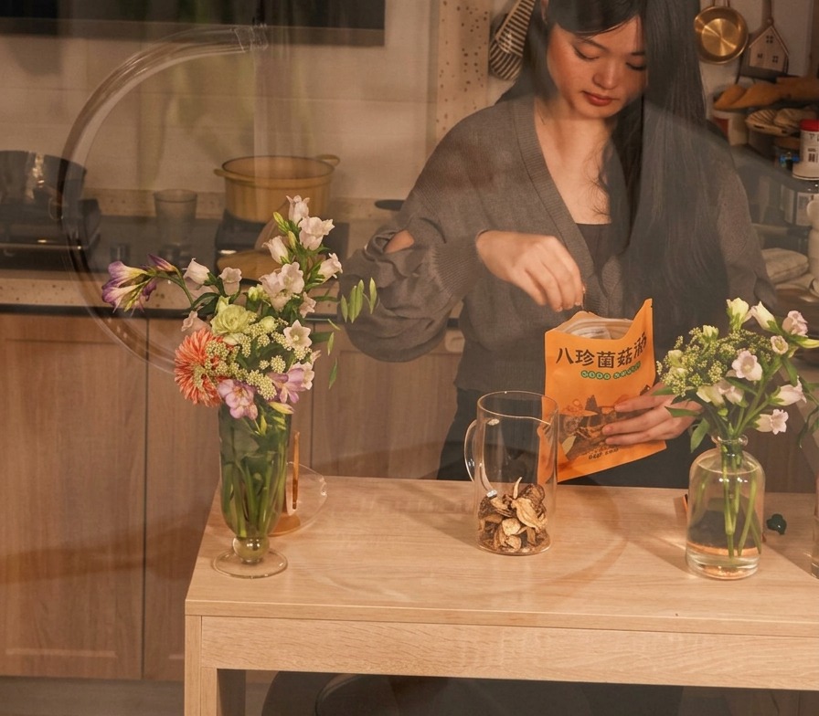 A person placing dried mushrooms from a bag into a clear glass pitcher on a wooden table.