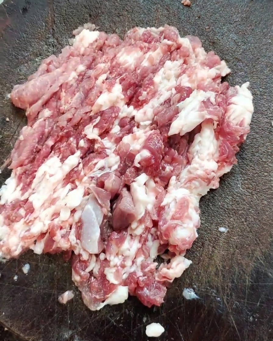 A sharp cleaver resting on a mound of freshly chopped pork shoulder meat on a textured wooden board.