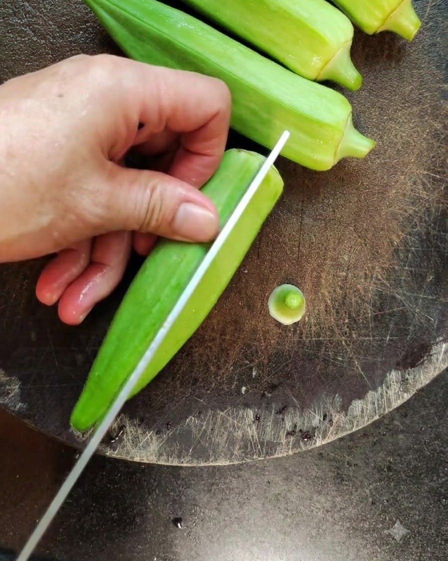 A close-up of a knife splitting a fresh green okra lengthwise on a dark cutting board.